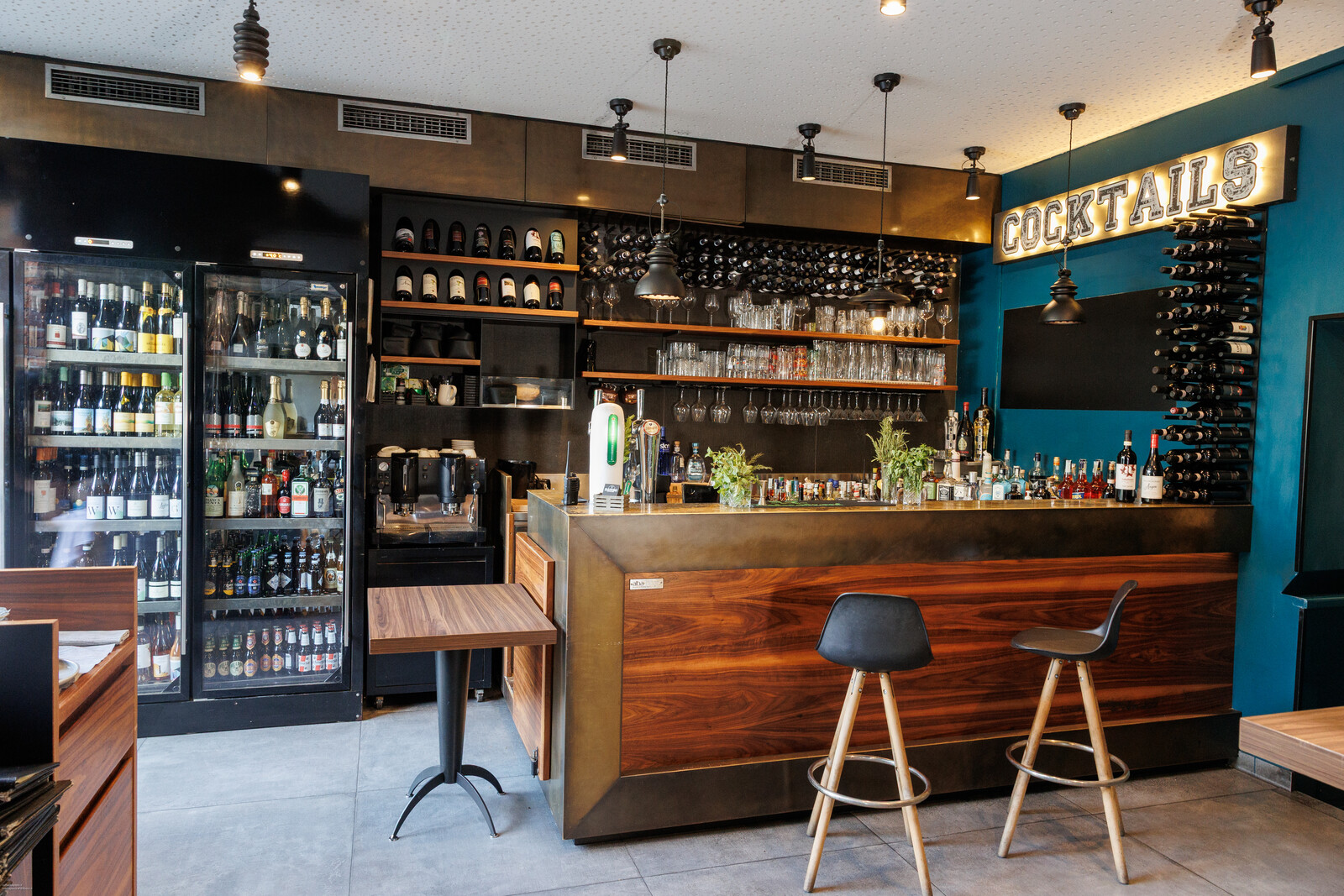Il bancone del bar con gli sgabelli, le vetrine dei vini e l'insegna Cocktails