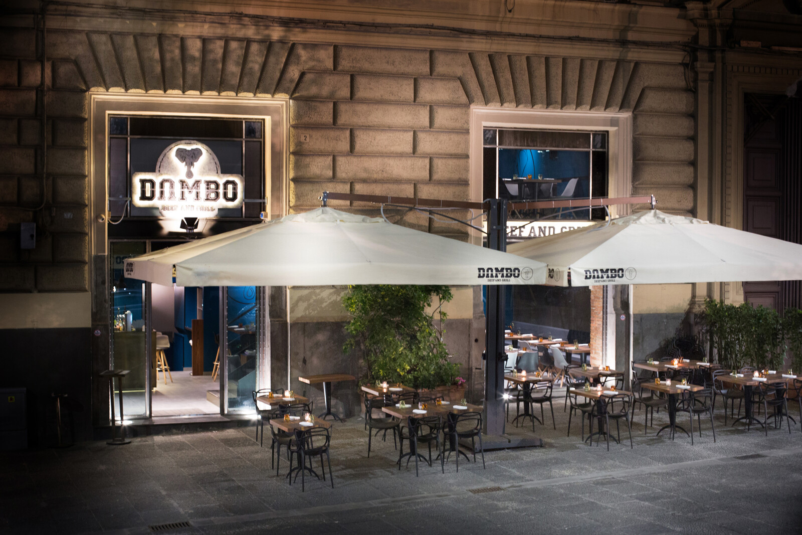 L'ingresso di Dambo di sera, con l'insegna illuminata, gli ombrelloni bianchi e i tavoli del dehors apparecchiati